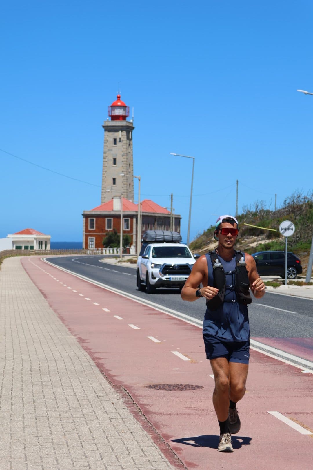 Jonathan Fors running towards the camera along a coastal road, a lighthouse and his support truck behind him.
